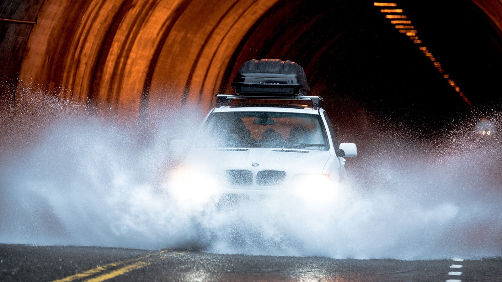 A silver BMW X5 with a roof box appears to aquaplane as it hits a large puddle while exiting a tunnel
